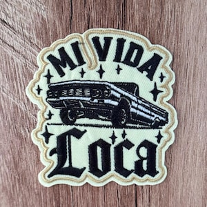 Könnte beinhalten: Ein bestickter Aufnäher mit einer schwarzen Lowrider-Auto-Illustration, zentriert zwischen dem Text MI VIDA in serifenloser Schrift und LOCA in gotischer Schrift. Das Design enthält kleine schwarze Sternakzente auf cremefarbenem Grund.