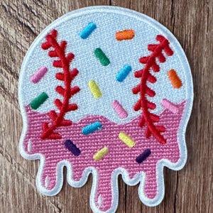 May include: A circular embroidered patch featuring a white baseball design with red stitching, partially covered by pink icing that drips down the bottom edge. The surface is decorated with small, colorful sprinkles in blue, green, orange, yellow, and purple.