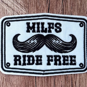 May include: A rectangular embroidered patch with a white background and a black border. The patch features the black embroidered text MILFS at the top and RIDE FREE at the bottom, with a large black mustache graphic centered between the two lines.