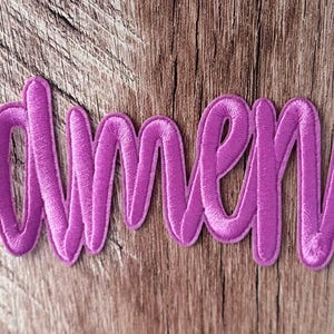 May include: A purple embroidered patch featuring the word amen in a cursive script font. The lettering has a raised, stitched texture with a consistent thread density throughout the design, creating a clear and legible word in a vibrant purple color.