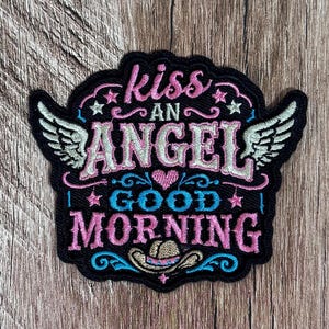 May include: A black embroidered patch featuring the text kiss AN ANGEL GOOD MORNING in pink, white, and blue thread. The design includes two white wings on the sides, a small pink heart, several stars, and a tan cowboy hat at the bottom.