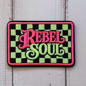 May include: A rectangular embroidered patch featuring a black and lime green checkered background. The text REBEL SOUL is stitched in the center, with REBEL in hot pink and SOUL in lime green. The entire patch is bordered by a hot pink stitched edge.