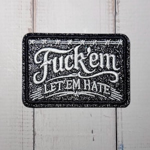 Könnte beinhalten: Ein rechteckiger bestickter Aufnäher mit schwarzem Glitzerhintergrund und weißem Rand. Der Text "Fuck'em" ist in einer großen, stilisierten weißen Schreibschrift gestickt, darunter stehen die Worte "LET EM HATE" in einer kleineren, fetten Schrift.