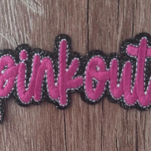 May include: A fabric patch featuring the word pinkout embroidered in bright pink thread. The lettering has a thick black border with white stitching details along the edges, creating a bold contrast against the vibrant pink script font.