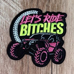 May include: An embroidered patch featuring a black off-road vehicle with pink accents and large tires. The text LET'S RIDE in pink and BITCHES in lime green is stitched above the vehicle, with a white tire tread graphic curving along the left side.