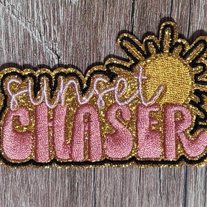 May include: An embroidered patch featuring a gold glitter sun graphic and the text "sunset CHASER" in light pink cursive and block lettering. The design is outlined in black thread, creating a bold, textured appearance with a shimmering finish.
