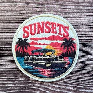 May include: A circular embroidered patch featuring a pontoon boat on water with palm trees and a sunset background. The word SUNSETS is stitched in red capital letters at the top. The design uses vibrant red, orange, yellow, blue, and black thread.