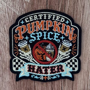 Puede incluir: Un parche bordado negro con el texto CERTIFIED PUMPKIN SPICE HATER. El diseño incluye dos manos de esqueleto sosteniendo tazas de café, un círculo de prohibición rojo sobre una calabaza y un café latte, y detalles a cuadros blancos y negros.