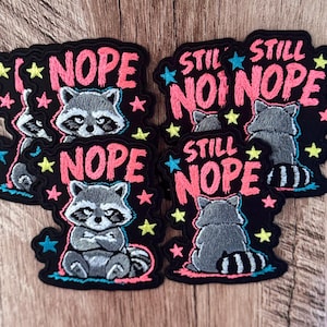 May include: Embroidered patches featuring a gray raccoon with a black mask and striped tail. Some patches show the raccoon facing forward with crossed arms and the text NOPE, while others show the raccoon from behind with the text STILL NOPE.