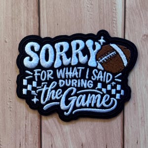 以下が含まれることがあります： SORRY FOR WHAT I SAID DURING the Gameという白い文字が刺繍された黒いワッペン。茶色のフットボールのグラフィックと両サイドの白い市松模様がアクセントになっており、全体が厚い黒い縁取りで囲まれ、ステッチ仕上げが施されています。