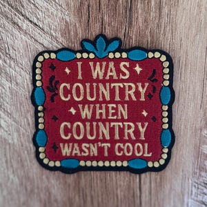 May include: A square embroidered patch with a red background and gold text that reads I WAS COUNTRY WHEN COUNTRY WASN'T COOL. The border is black with gold dots and teal accents, featuring a decorative top element and small black star motifs.