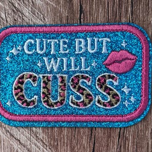 May include: A rectangular embroidered patch with a bright blue glitter background and a pink border. The text reads CUTE BUT WILL CUSS in white and leopard print lettering. A small pink lip icon and several small white star accents are included.