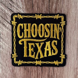 May include: A square embroidered patch with a black background and gold thread. The text reads "CHOOSIN' TEXAS" in a bold, stylized font. The border is designed to look like barbed wire, also rendered in gold embroidery thread.