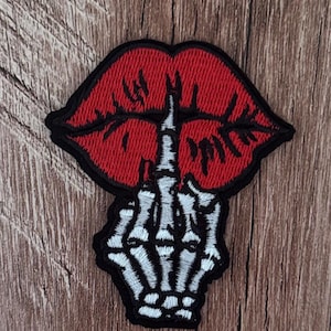 May include: An embroidered patch featuring a pair of bright red lips with a black outline. A white and grey skeleton hand is positioned vertically in front of the lips, with the index finger raised in a shushing gesture against the mouth.