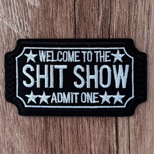 May include: A rectangular black embroidered patch with a white border shaped like a ticket. The text reads WELCOME TO THE SHIT SHOW and ADMIT ONE, accented by four white stars. The design mimics a vintage admission ticket style.