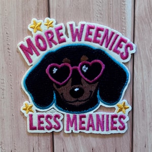 Puede incluir: Un parche bordado que muestra un perro salchicha negro con gafas de sol rosas en forma de corazón. El texto MORE WEENIES LESS MEANIES aparece en letras rosas, decorado con pequeñas estrellas amarillas sobre un fondo blanco con borde azul.