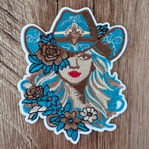 May include: An embroidered patch featuring a person wearing a bright blue cowboy hat with brown accents. The design includes flowing hair, a face with red lips, and a cluster of brown and blue flowers surrounding the lower portion of the image.