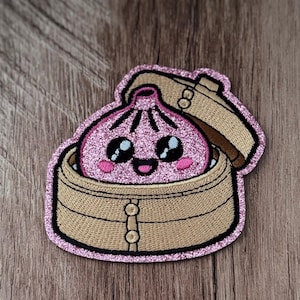 May include: An embroidered patch featuring a pink glittery dumpling with a smiling face inside a tan bamboo steamer basket. The design has a thick pink glitter border, black embroidered outlines, and small pink blush marks on the dumpling cheeks.