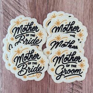Puede incluir: Una colección de parches bordados de color crema con bordes festoneados. Varios parches presentan el texto negro Mother of the Bride con detalles de estrellas doradas, mientras que un parche presenta el texto negro Mother of the Groom con un corazón dorado.