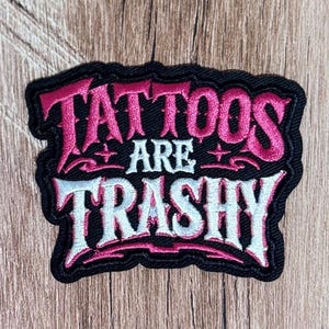 May include: An embroidered patch with a black border featuring the text TATTOOS ARE TRASHY. The word TATTOOS is in pink, the word ARE is in white, and the word TRASHY is in white with pink outlines, all set against a black background.