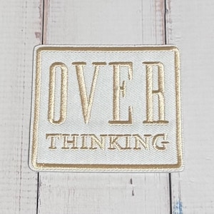 May include: A rectangular embroidered patch featuring a white background with a gold border. The word OVER is stitched in large gold capital letters above the word THINKING, which is also stitched in gold capital letters on the white fabric.