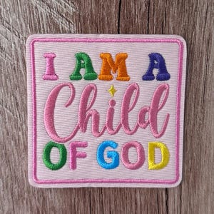 Könnte beinhalten: Ein quadratischer gestickter Aufnäher mit hellrosa Hintergrund und rosa Rand. Der Text lautet I AM A Child OF GOD in mehrfarbiger Block- und Schreibschrift. Ein kleiner gelber Stern befindet sich über dem Wort Child in der Mitte.