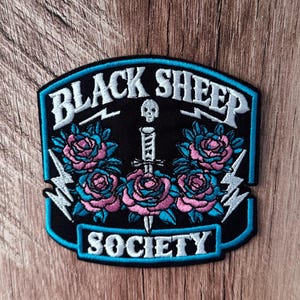 May include: An embroidered patch with a black background featuring the text "BLACK SHEEP" at the top and "SOCIETY" at the bottom in white. The design includes a central white dagger with a skull, surrounded by pink and blue roses and lightning bolts.