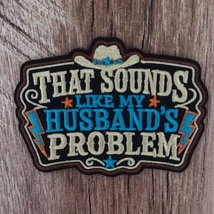 May include: A black embroidered patch featuring a white cowboy hat at the top and the text THAT SOUNDS LIKE MY HUSBAND'S PROBLEM in white and blue lettering. The design includes two orange stars, a blue star, and two blue lightning bolts.