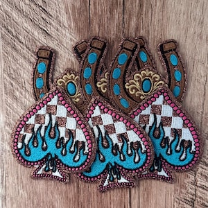 May include: A collection of embroidered patches featuring a spade shape with a white and brown checkered pattern, topped with blue dripping flames and outlined in pink dots. Behind these are brown horseshoes with blue accents and gold details.