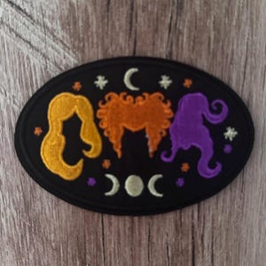 May include: An oval-shaped black embroidered patch featuring three stylized hair silhouettes in yellow, orange, and purple. The design includes small white stars, a crescent moon at the top, and a triple moon phase symbol at the bottom.