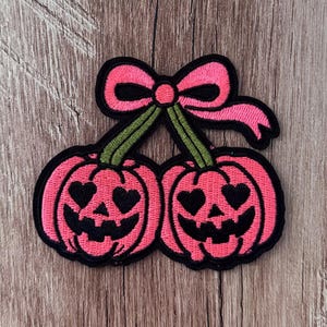May include: An embroidered patch featuring two pink pumpkins with heart-shaped eyes and black jagged smiles. The pumpkins are connected by green stems topped with a pink bow, all outlined in black thread against a textured wooden surface.