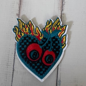 Puede incluir: Un parche bordado con forma de corazón con un patrón de cuadros en color verde azulado y negro. El corazón está rematado con llamas amarillas y rojas y presenta dos esferas rojas similares a cerezas, cada una con un ocho negro en el centro.
