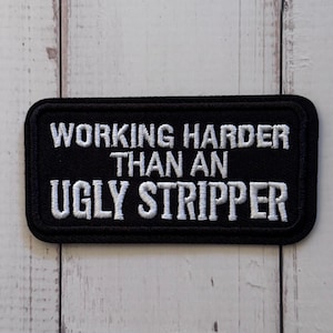 May include: A rectangular black embroidered patch with rounded corners featuring the white stitched text "WORKING HARDER THAN AN UGLY STRIPPER" in a bold, sans-serif font. The patch has a thick black border and a textured fabric finish.