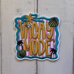 Puede incluir: Un parche bordado con un borde ondulado color cerceta sobre un fondo de rayas verticales rosas y blancas. El texto VACAY MODE está bordado en hilo dorado. Los elementos decorativos incluyen una estrella de mar amarilla, dos conchas verdes y dos palmeras negras.