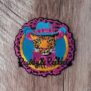 May include: An embroidered patch featuring a leopard wearing a bright blue cowboy hat. The leopard has yellow fur with black spots and blue eyes. The design includes a pink bow, a yellow circle, and a pink scalloped border with the text Rowdy & Reckless.