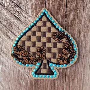 May include: An embroidered spade-shaped patch featuring a brown and tan checkered pattern. The design is accented with dark brown floral embroidery along the sides and is bordered by a continuous line of bright turquoise circular stitching.