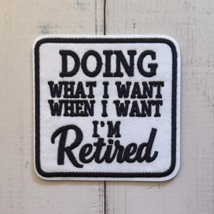 May include: A square embroidered patch with a white background and a thick black border. The text in black embroidery reads: DOING WHAT I WANT WHEN I WANT I'M Retired. The patch features a clean, bold font for the first three lines and a script font.
