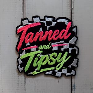 Puede incluir: Un parche bordado rectangular con un borde de cuadros blancos y negros. El texto Tanned en rosa brillante y Tipsy en verde neón está cosido en una fuente cursiva en el centro, con la palabra and en un texto verde más pequeño.