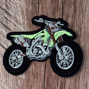 Può includere: Una toppa ricamata a forma di moto da cross. Il design presenta un telaio e una sella di colore verde lime brillante, con dettagli del motore in grigio e bianco, accenti marroni e pneumatici neri. L'intera moto è delineata da un bordo nero.