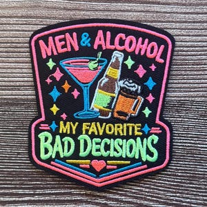 May include: A black embroidered patch with a shield shape featuring the text MEN & ALCOHOL MY FAVORITE BAD DECISIONS in pink, yellow, and green. The design includes a pink martini glass, a beer bottle, and a beer mug with colorful star accents.