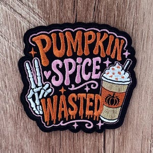 May include: An embroidered patch with a black background featuring the orange and pink text PUMPKIN SPICE WASTED. The design includes a white skeleton hand making a peace sign, a coffee cup with whipped cream, and small pink star accents.