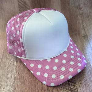 May include: A trucker hat featuring a solid white foam front panel and a pink mesh back panel. The pink brim and mesh back are decorated with a white polka dot pattern. A small pink button sits at the top center of the crown, and a white cord trim.
