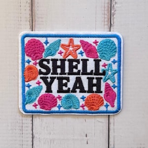 May include: A rectangular embroidered patch with a white background and a blue border. The text SHELL YEAH is stitched in black in the centre, surrounded by colourful embroidered seashells and starfish in shades of pink, orange, and teal.