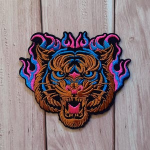 May include: An embroidered patch featuring a tiger head with an open mouth, rendered in shades of brown, black, and bright pink, surrounded by stylised blue and pink flames. The design is outlined in black thread on a dark fabric base.