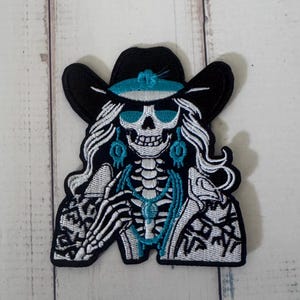 Puede incluir: Un parche bordado que muestra un esqueleto con un sombrero de vaquero negro con una banda turquesa, gafas de sol turquesas y pendientes y collar a juego. El esqueleto tiene el pelo largo y blanco y viste una chaqueta blanca estampada.