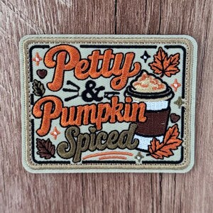 Peut inclure: Un écusson brodé rectangulaire avec un fond vert clair et une bordure beige. Il présente le texte "Petty & Pumpkin Spiced" en écriture orange et vert olive. Une illustration d'une tasse de café avec de la crème fouettée et des feuilles.