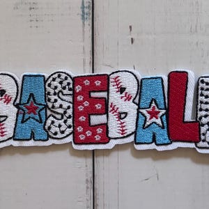 May include: An embroidered patch featuring the word BASEBALL in block letters. The letters B, S, and L have a white baseball texture with red stitching. The A and A are light blue with red stars. The E is white with black dots, and the B is red.