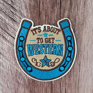 May include: An embroidered patch shaped like a horseshoe in bright blue and tan. The center features the text IT'S ABOUT TO GET WESTERN in brown and blue, accented with two blue stars and decorative swirls on a tan background.