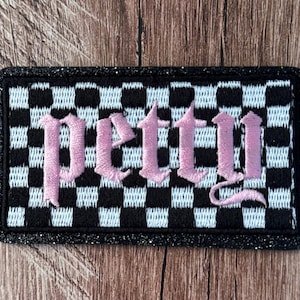 May include: A rectangular embroidered patch featuring a black and light blue checkered pattern background. The word petty is stitched across the center in a light pink gothic-style font. The patch has a thick, black, glittery border.