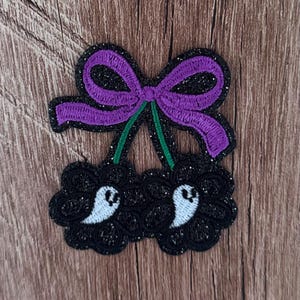 May include: An embroidered patch featuring two black glittery cherries with white ghost faces on each fruit. The cherries are connected by green stems to a purple embroidered bow at the top. The entire design is outlined in black glitter thread.